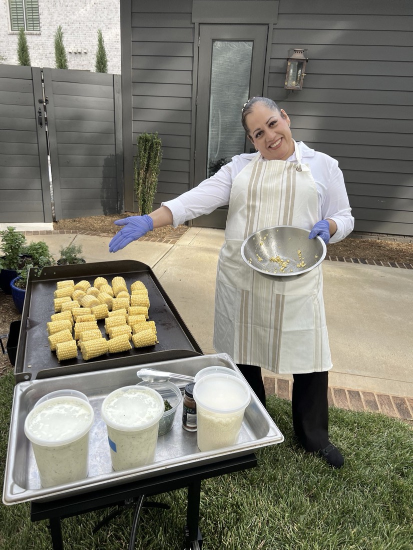 Alba serving at an outdoor party, beside a griddle of fresh corn for a live elote station